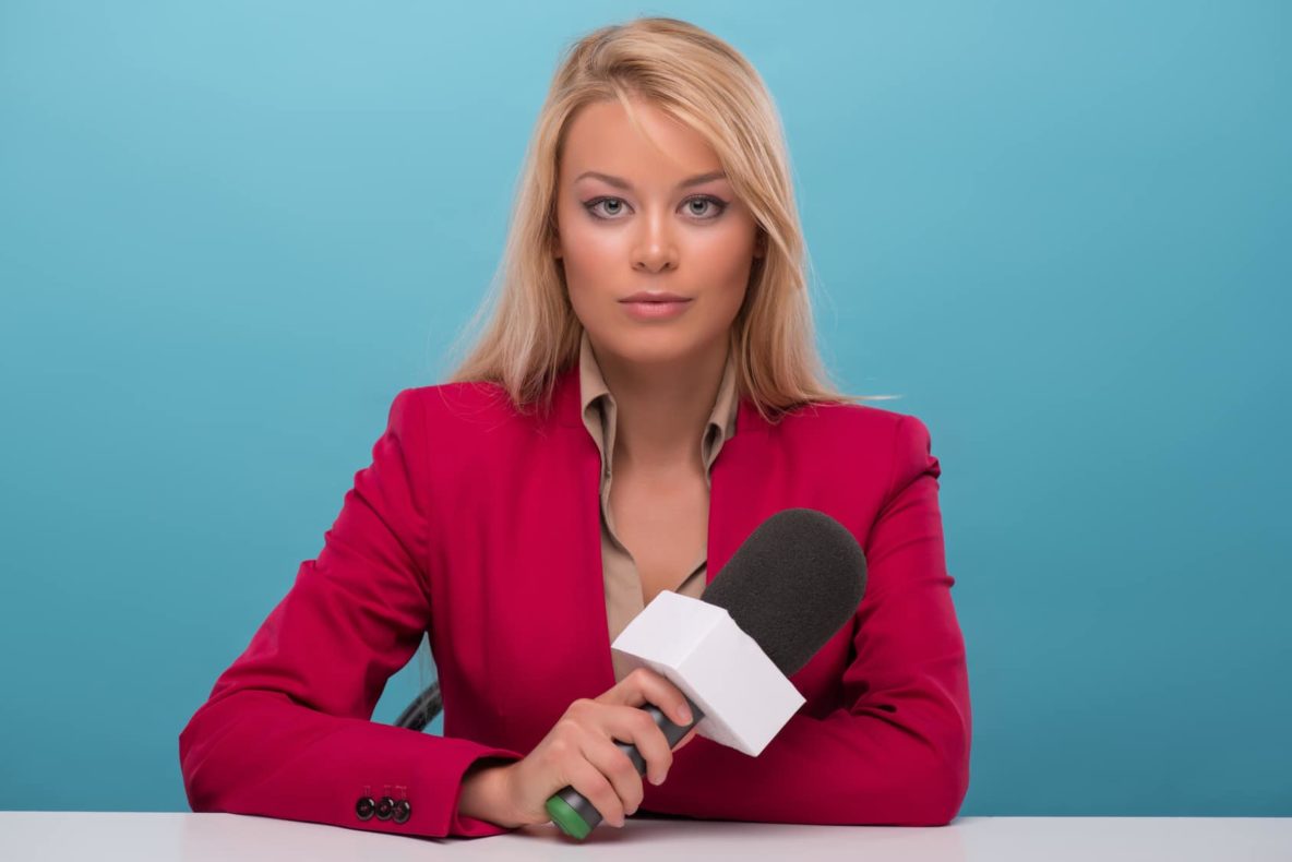 Half-length portrait of good-looking fair-haired TV presenter wearing great red jacket and cream-colored shirt sitting at the table holding a microphone. Isolated on blue background